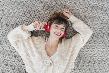 Happy woman listening music through wireless headphones lying on carpet at home - PNAF05062