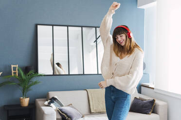 Unbeschwerte Frau mit Kopfhörern tanzt mit erhobener Hand im Wohnzimmer zu Hause - PNAF05067