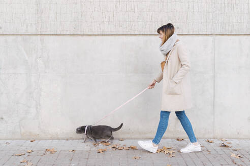 Frau mit Hauskatze auf dem Gehweg an einer Mauer spazierend - PNAF05090