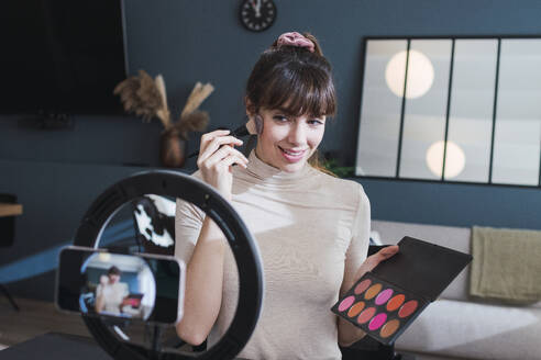 Lächelnde Frau beim Filmen eines Make-up-Vlogs mit Palette zu Hause - PNAF05109