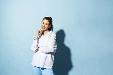 Happy young woman standing with hand on chin against blue background - MIKF00231