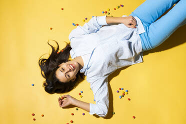 Glückliche junge Frau inmitten von bunten Bonbons gelben Hintergrund liegen - MIKF00237