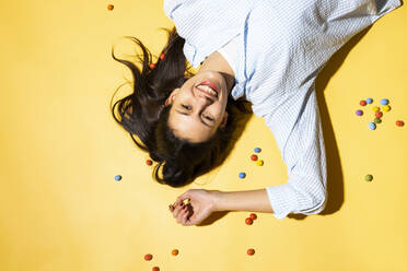 Glückliche junge Frau inmitten von bunten Bonbons vor farbigem Hintergrund liegend - MIKF00238