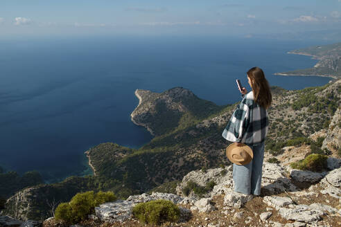 Frau, die auf einer Klippe stehend das Meer mit ihrem Smartphone fotografiert - SYEF00317