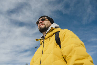 Happy man wearing sunglasses standing under cloudy sky - ALKF00186