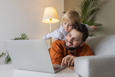 Happy father using laptop with son lying on father at home - AAZF00164