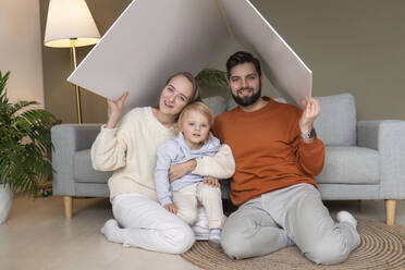 Happy father and mother with son sitting under cardboard roof at home - AAZF00169