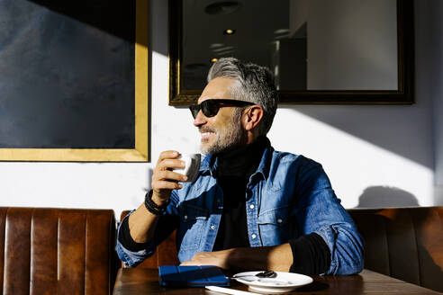 Lächelnder reifer Geschäftsmann mit Kaffeetasse in einem Café sitzend - JJF00689