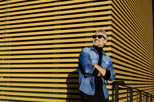 Mature man standing with arms crossed in front of yellow striped building - JJF00701