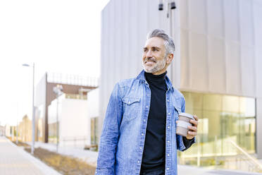 Smiling mature man with coffee cup standing in front of building - JJF00721