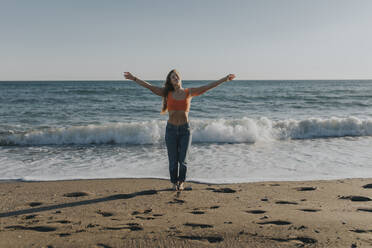 Carefree woman standing with arms outstretched in front of sea at beach - DMGF01103