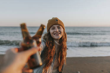 Glückliche Frau stößt mit einem Freund vor dem Meer auf eine Bierflasche an - DMGF01106