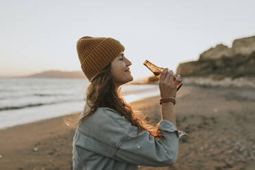 Smiling woman with beer bottle standing at beach - DMGF01108