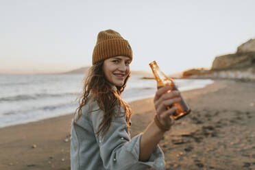 Happy woman with beer bottle standing at beach - DMGF01109