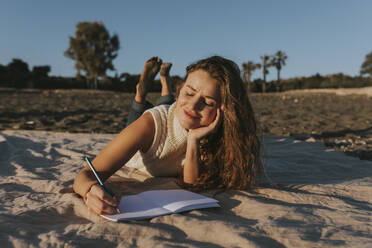 Smiling woman writing on note pad and lying at beach - DMGF01111