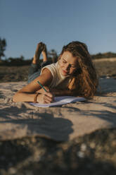 Woman writing on note pad and lying at beach - DMGF01112
