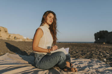 Nachdenkliche Frau sitzt mit Notizblock am Strand - DMGF01113