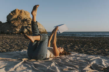 Woman with note pad lying on picnic blanket at beach - DMGF01114