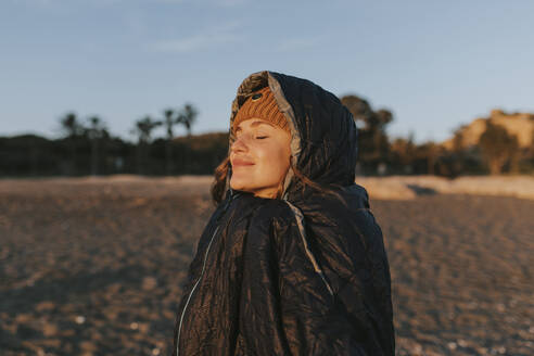 Smiling woman with eyes closed in sleeping bag at beach - DMGF01118