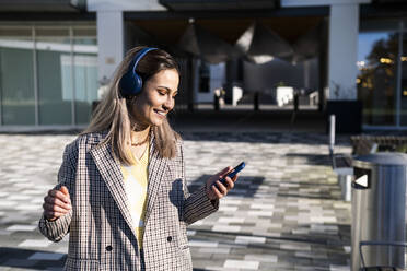 Lächelnde Geschäftsfrau mit drahtlosen Kopfhörern und Smartphone in der Hand an einem sonnigen Tag - AMWF01261