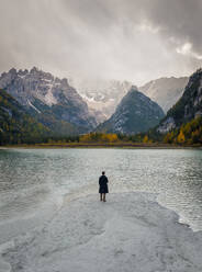 Drone view of anonymous traveler standing on shore near lake and admiring picturesque mountains in Dolomites Mountain ridge - ADSF43433