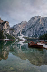 Landschaftlicher Blick auf einen ruhigen See mit Booten, in denen sich verschneite Berge mit Nadelbäumen spiegeln, an einem Wintertag in den Dolomiten - ADSF43436