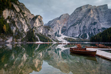 Landschaftlicher Blick auf einen ruhigen See mit Booten, in denen sich verschneite Berge mit Nadelbäumen spiegeln, an einem Wintertag in den Dolomiten - ADSF43437