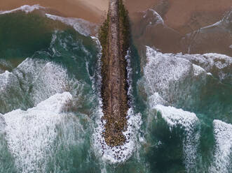 Aerial view of a majestic breakwater with rough rolling water waves rolling on the beach at Costa da Caparica, Setubal, Portugal. - AAEF17680