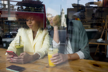 Frau, die ihr Smartphone mit einem Freund in einem Café teilt, gesehen durch ein Fenster - MMPF00702