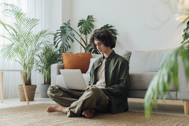 Freelancer using laptop sitting on carpet in front of sofa at home - AAZF00230