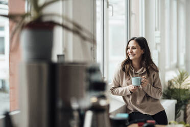 Glückliche Frau mit Kaffeetasse schaut durch ein Fenster - JOSEF17659