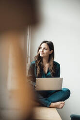 Nachdenkliche Geschäftsfrau sitzt mit Laptop auf der Fensterbank im Büro zu Hause - JOSEF17660
