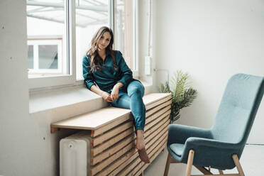 Confident young businesswoman sitting by windowsill in office - JOSEF17666