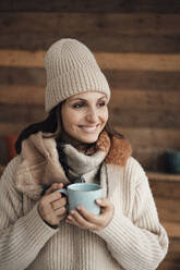 Glückliche Frau in warmer Kleidung mit Kaffeetasse im Stehen zu Hause - JOSEF17683