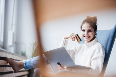 Happy freelancer holding credit card sitting with laptop in chair at home office - JOSEF17699