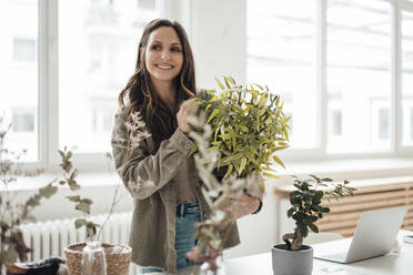 Happy businesswoman standing with potted plant at home office - JOSEF17702