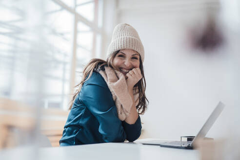 Happy businesswoman wearing knit hat sitting in front of laptop at home office - JOSEF17757