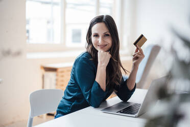 Lächelnde Geschäftsfrau, die eine Kreditkarte hält, sitzt am Tisch im Büro zu Hause - JOSEF17760