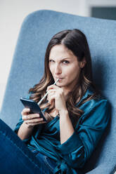 Young woman holding straw on smart phone sitting in armchair - JOSEF17765