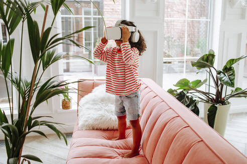 Girl wearing virtual reality simulator standing on sofa in living room at home - MDOF00903