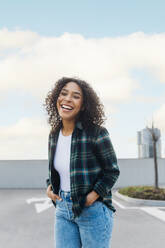 Cheerful young woman with curly hair standing on footpath - MEUF08945