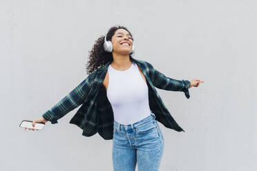 Happy young woman wearing wireless headphones dancing in front of wall - MEUF08955