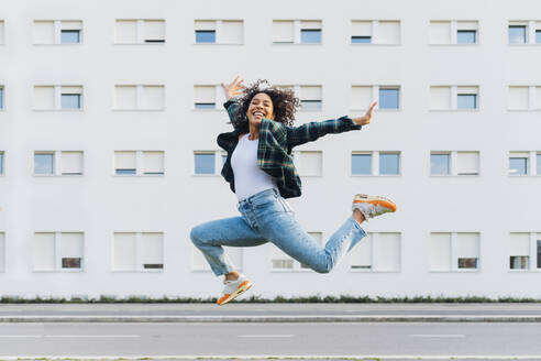 Cheerful young woman jumping in front of building - MEUF08964