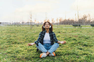 Junge Frau mit drahtlosen Kopfhörern beim Meditieren im Park - MEUF09012