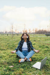 Young woman wearing wireless headphones meditating in park at sunset - MEUF09013
