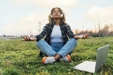 Woman wearing wireless headphones meditating in park at sunset - MEUF09014