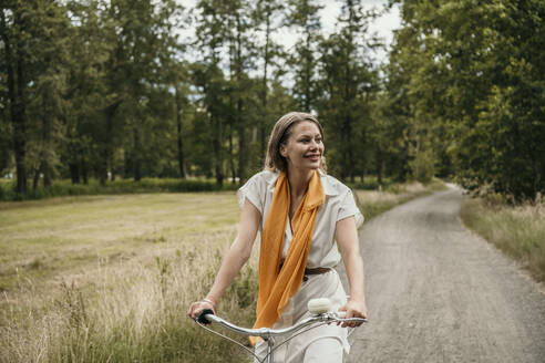 Nachdenklich lächelnde Frau fährt Fahrrad auf unbefestigtem Weg - MJRF00930