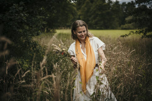 Smiling woman holding wildflowers in field - MJRF00935