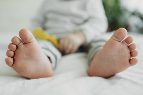 Barefoot feet of toddler boy sitting on bed - ANAF01114