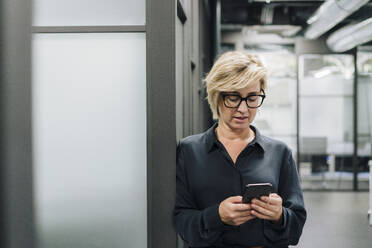 Blond businesswoman using mobile phone leaning on wall at office - JOSEF17822